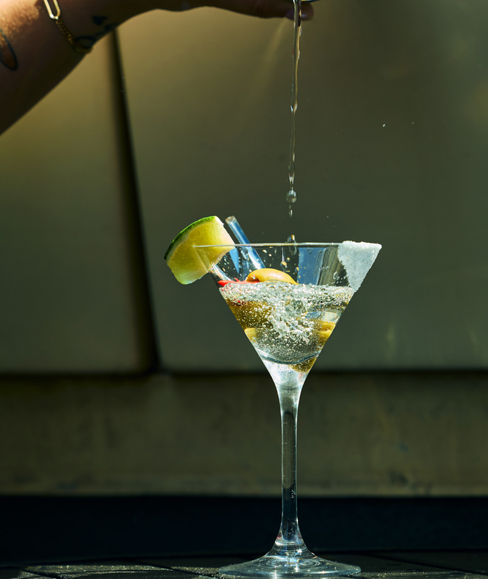 Hand pouring a clear liquid into a martini glass garnished with a lime wedge and olives.