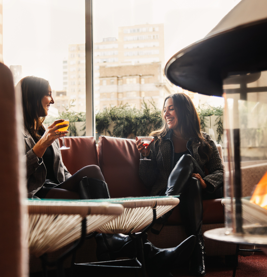 Two women enjoy drinks and conversation by a patio fire on a cozy, sunlit day.