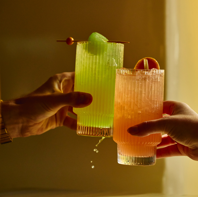 Two hands clink glasses filled with colorful drinks, one green with a lemon slice, one orange with a citrus slice, against a warm background.