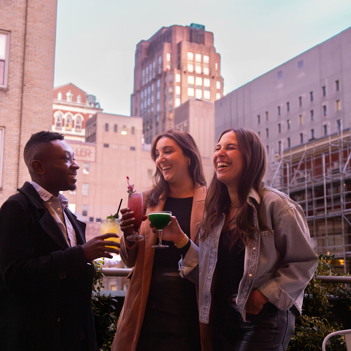 Three people enjoy drinks on a rooftop, smiling and laughing against a cityscape backdrop.