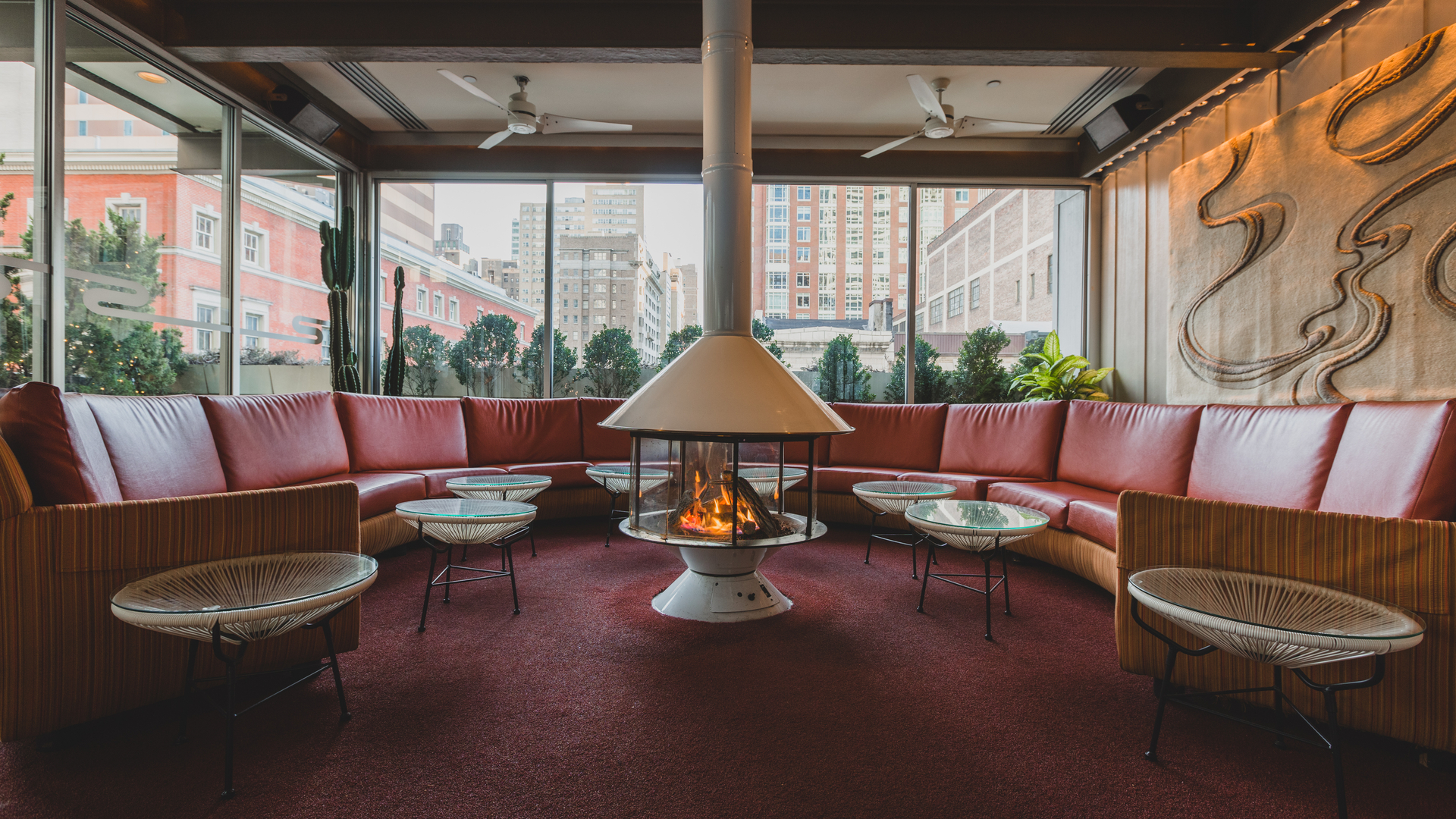 Modern restaurant lounge with sectional red couches, fire pit, patterned chairs, and large windows overlooking city buildings.