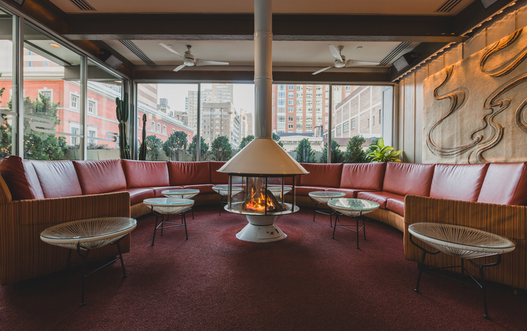 Sofa seating around a central fireplace in a cozy restaurant interior with large windows and red carpet.