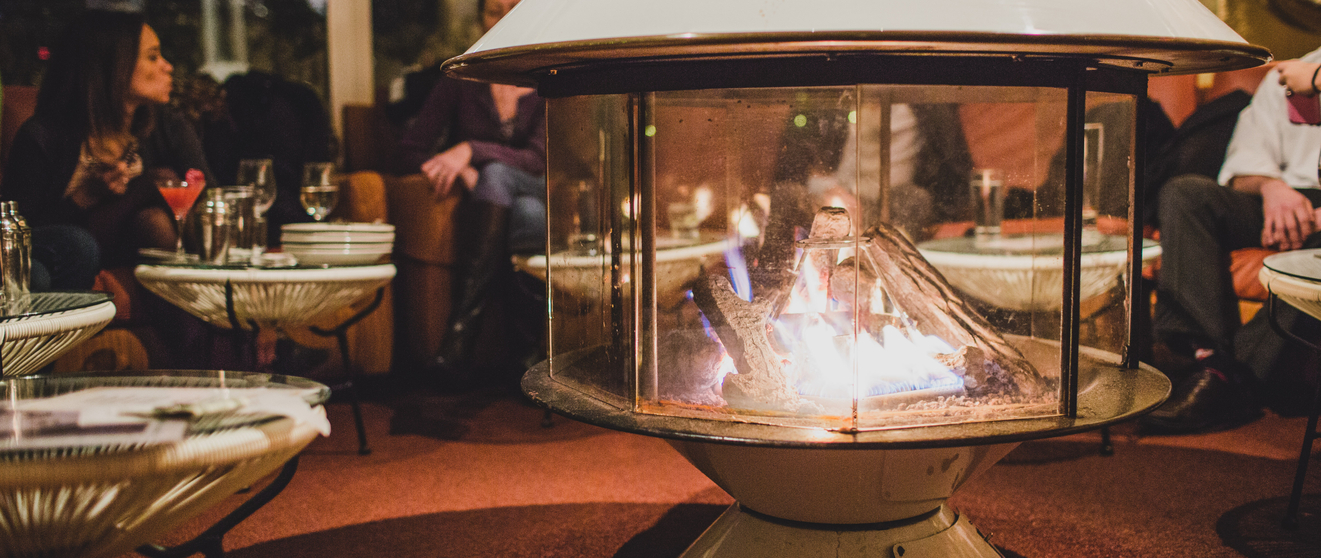 Restaurant interior with people dining, central gas fireplace, modern tables.