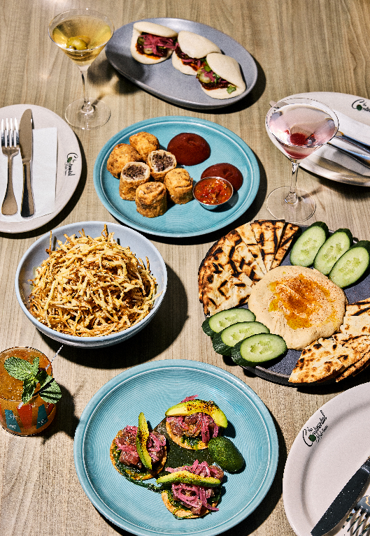 Overhead view of a varied restaurant spread on blue and grey plates, including sliced steak with avocado, crispy shoestring fries, stuffed mushrooms with dipping sauces, flatbread with hummus and cucumber, and bao buns, alongside wine and a cocktail.
