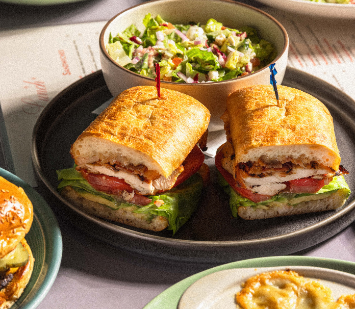 A toasted ciabatta chicken sandwich sliced in half and served on a dark plate alongside a chopped salad with red onion and greens in a white bowl.