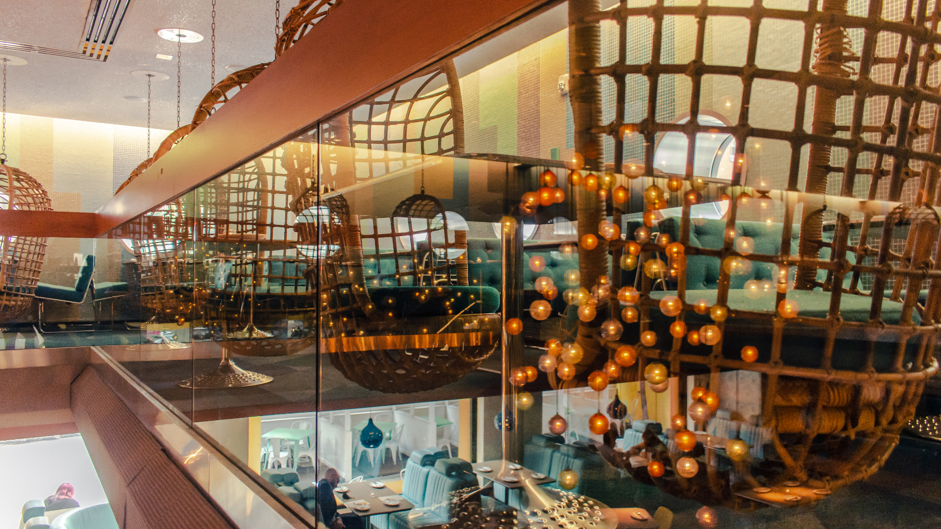 A dramatic multi-level restaurant interior viewed through glass, featuring hanging rattan egg chairs, cascading globe string lights, woven ceiling installations, and teal upholstered seating below.