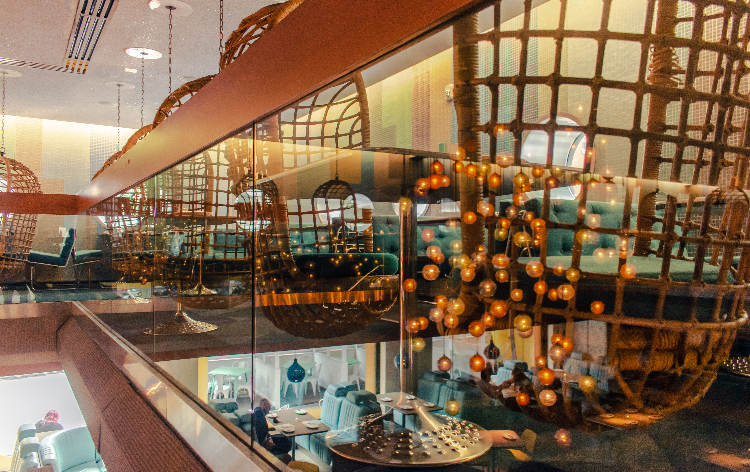 A dramatic multi-level restaurant interior viewed through glass, featuring hanging rattan egg chairs, cascading globe string lights, woven ceiling installations, and teal upholstered seating below.
